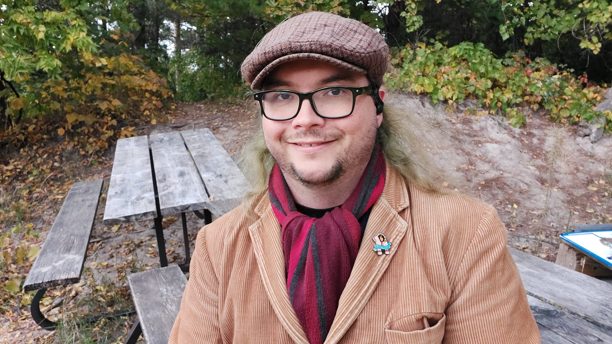 A man with long green hair sits on top of a picnic table, a forest path behind him